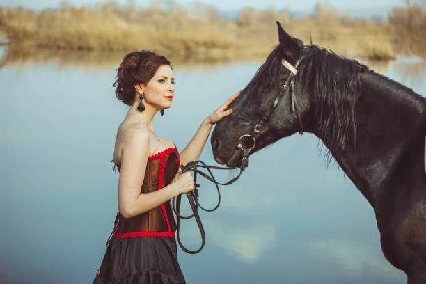 Black Corset: A Necessary Return for Breast Support? 1 Woman in black corset ensemble gently petting majestic dark horse by water.